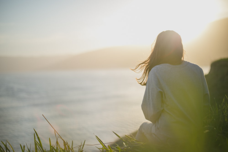 A person sitting on grassy ground, overlooking a body of water, embodying a moment of peaceful solitude resonating with the themes of trauma-informed therapy services.