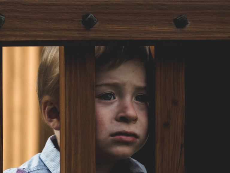 A young child with a sad expression looking out through a wooden fence.