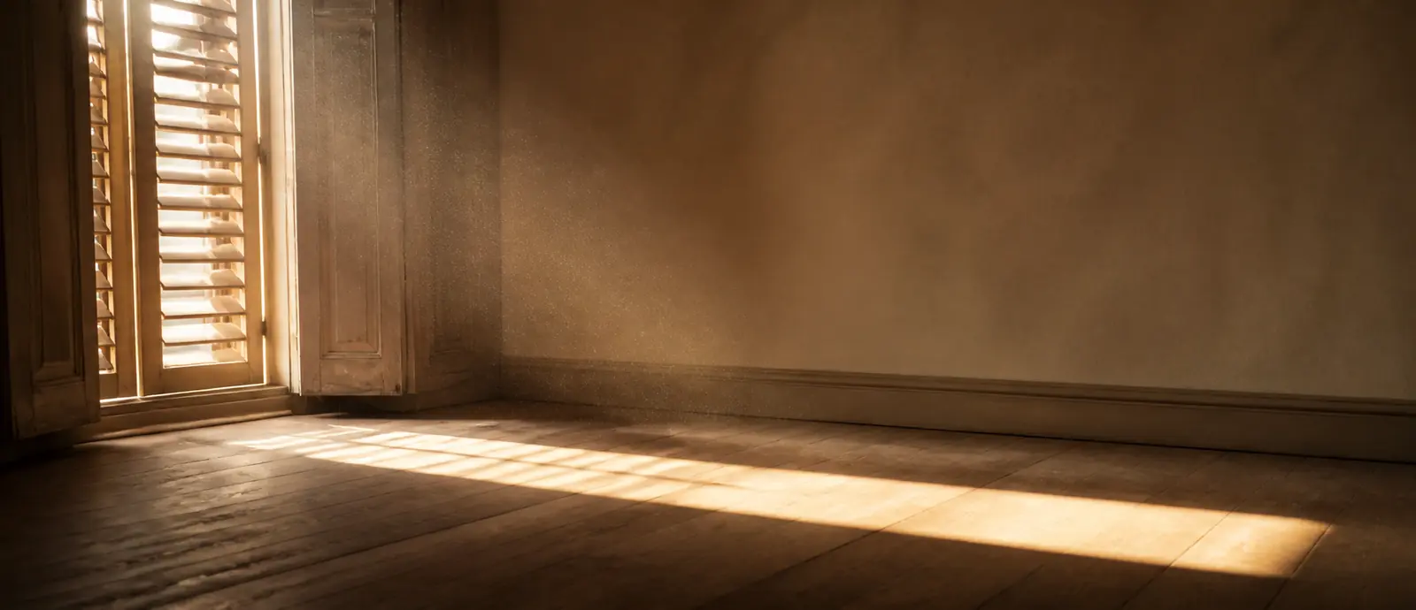 An empty room where afternoon light falls through a shuttered window onto a bare floor.